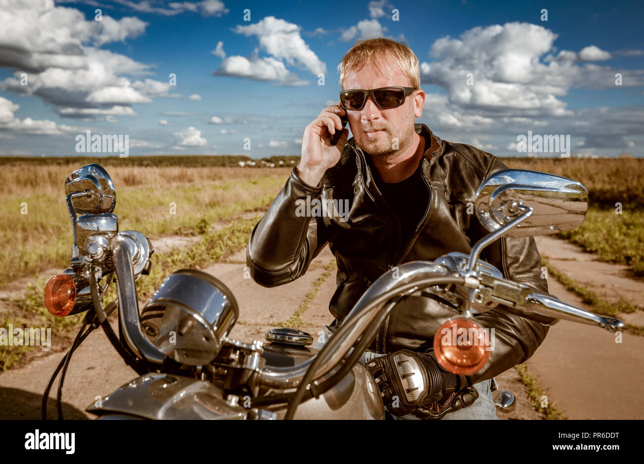 Biker parlando su uno smartphone. Biker uomo che indossa una giacca di pelle e occhiali da sole seduto sulla sua moto. Foto Stock