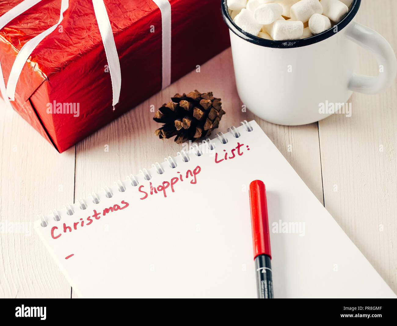 Regali di natale di pianificazione commerciale. Fare shopping o elenco di cose da fare per il Natale. Notebook, la tazza di cioccolata calda con marshmallows, regalo per l'anno nuovo e cono di pino bianco su sfondo di legno. Foto Stock