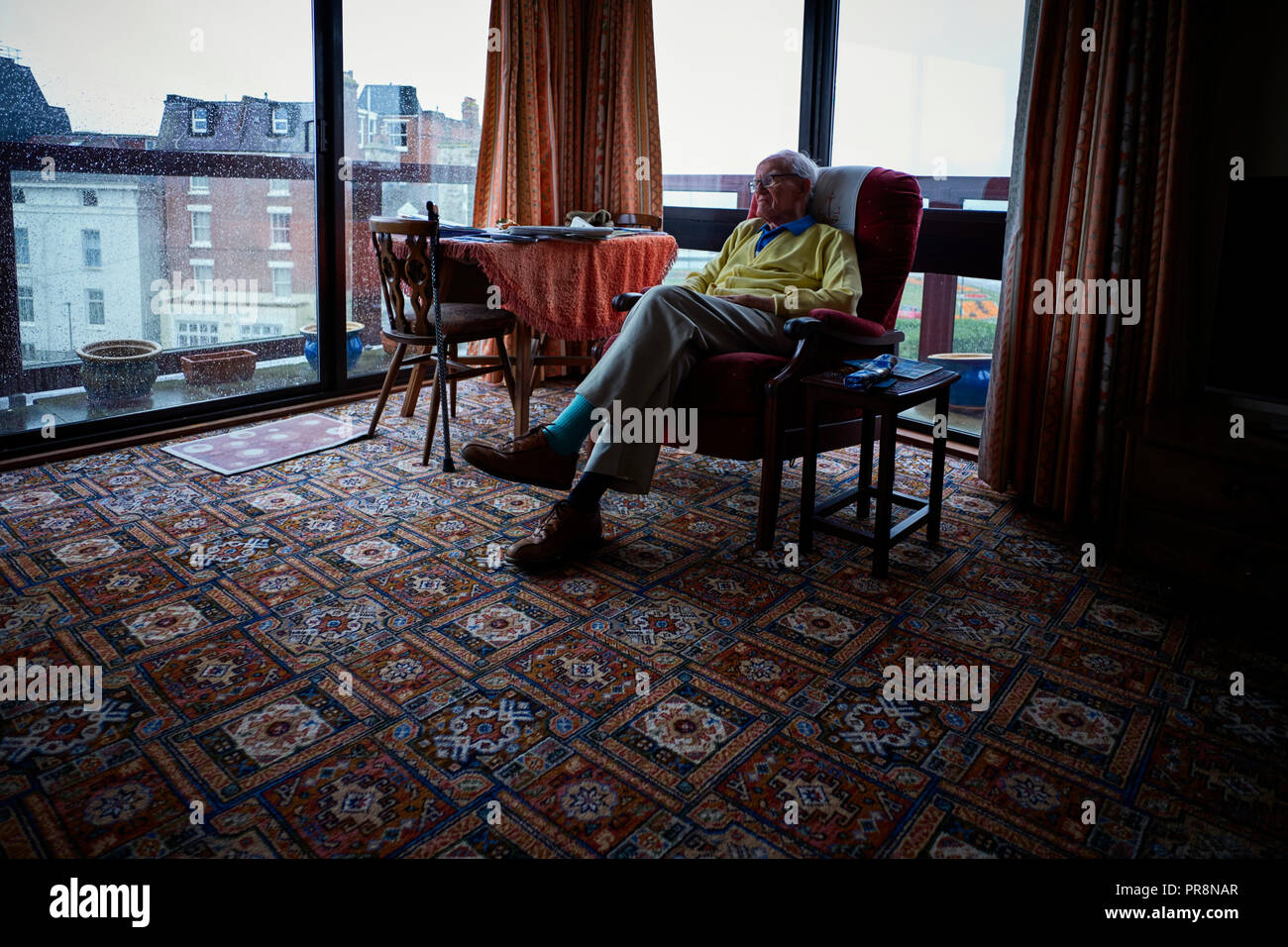I vecchi vedovo (87) seduto da solo nel suo appartamento sul mare con la pioggia battente esterno con la sua sedia girata in dalla vista mare Foto Stock