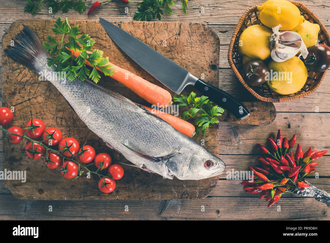 Shi drum pesce (Umbrina cirrosa) su una vecchia tavola di legno con carote, pomodori ciliegini, pomodori nero, limone, aglio, prezzemolo e peperoncino piccante, vista dall'alto s Foto Stock
