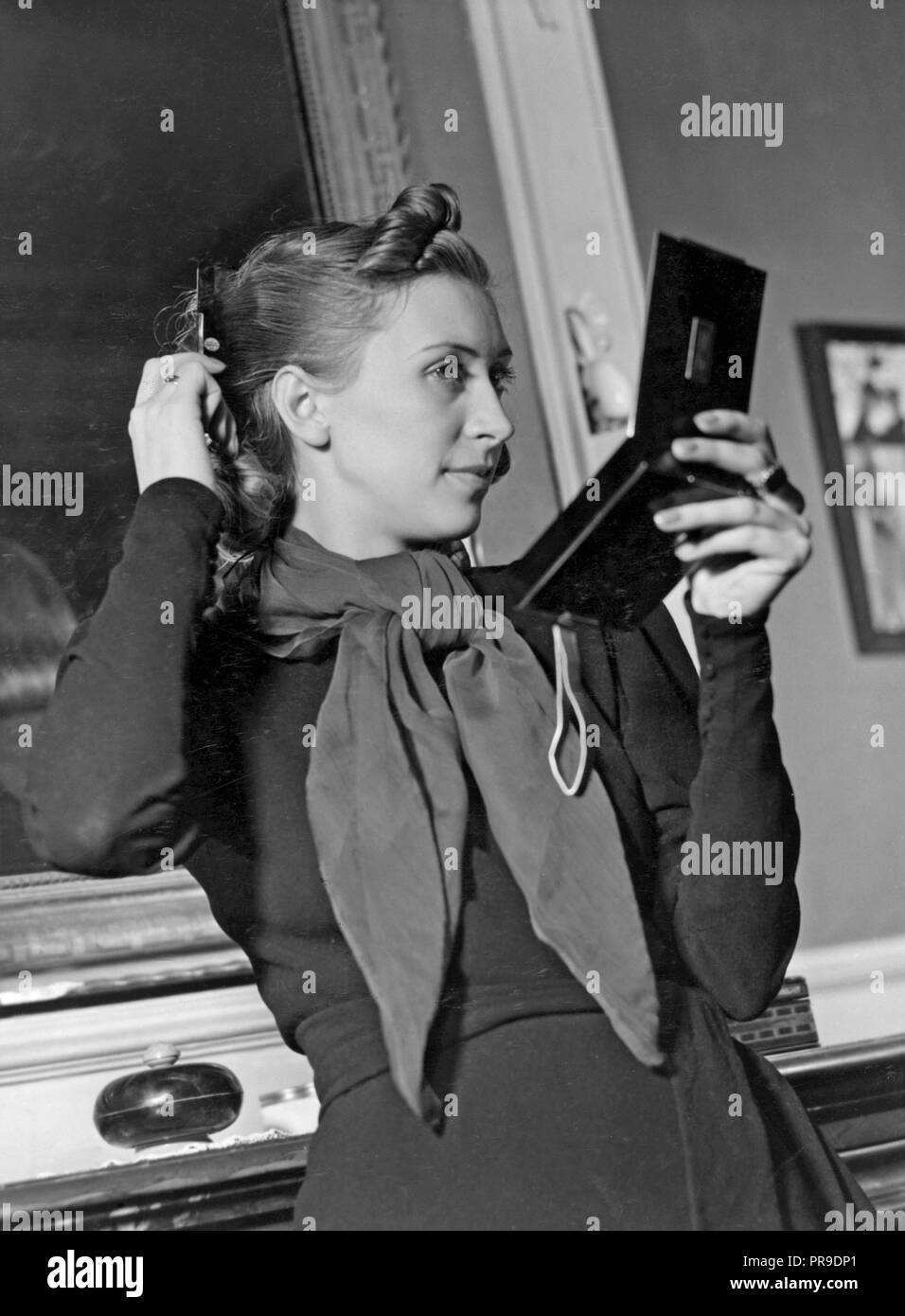 Donna pettinare i capelli. Un pelo scuro giovane donna è alla ricerca di sé in uno specchio tascabile mentre che fissa i suoi capelli. 1940s Foto Stock