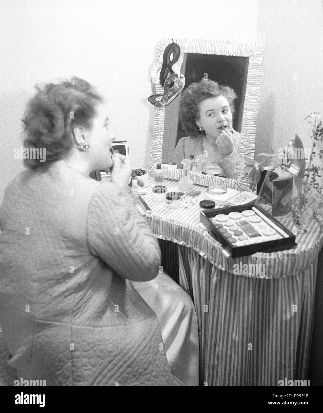 Cosmetici negli anni '1940s. Una giovane donna a casa sta sedendosi ad una scrivania di trucco e guardando se stessa nello specchio mentre si mette il rossetto. Ci sono molti tipi differenti di cosmettici che si trovano sul tavolo di fronte a lei. Svezia 1947. Foto Kristoffersson Ab3-8 Foto Stock
