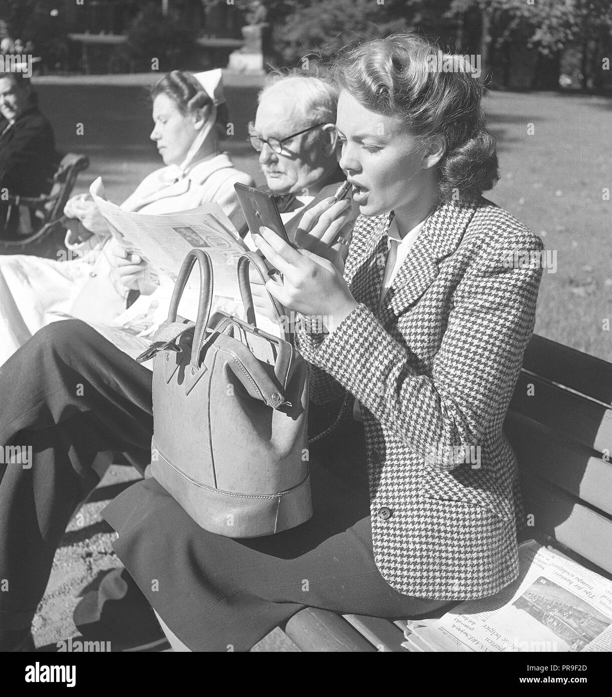 Fare fino a 1940s. La giovane attrice e il modello Haide Göransson, 1928-2008, sta mettendo il rossetto. Lei è seduta su una panchina di una giornata di sole in un parco e accanto a lei siede un anziano gentiluomo e un infermiere in uniforme, della maglieria. Haide ha la borsetta sul suo ginocchio e detiene un specchio tascabile in una mano quando si mette il rossetto. La Svezia 1949. Foto Kristoffersson ref A7-10 Foto Stock