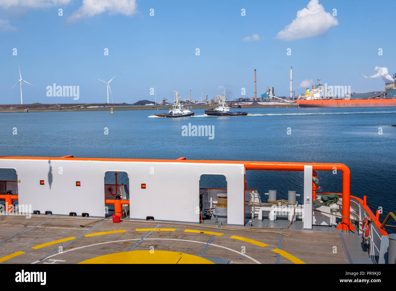 Vista di due rimorchiatori, le turbine eoliche e la fabbrica di acciaio dal ponte di una nave arancione nel porto di IJmuiden, Paesi Bassi. Foto Stock