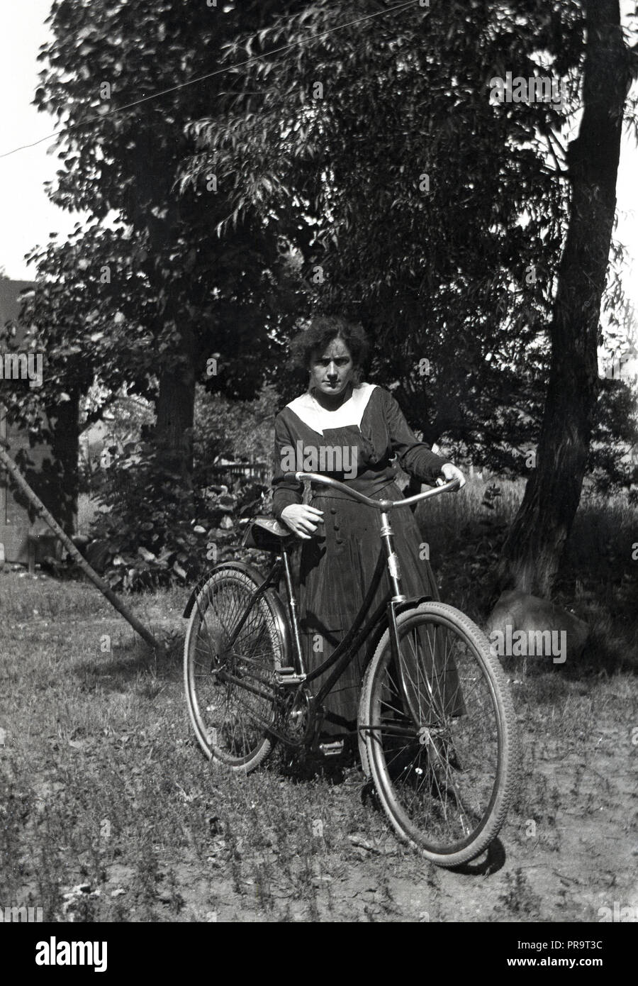 1920s, storico, una giovane donna che indossa un alto-collo abito lungo con maniche, comune a thiis era, in piedi fuori in una zona erbosa con la sua bicicletta. Foto Stock