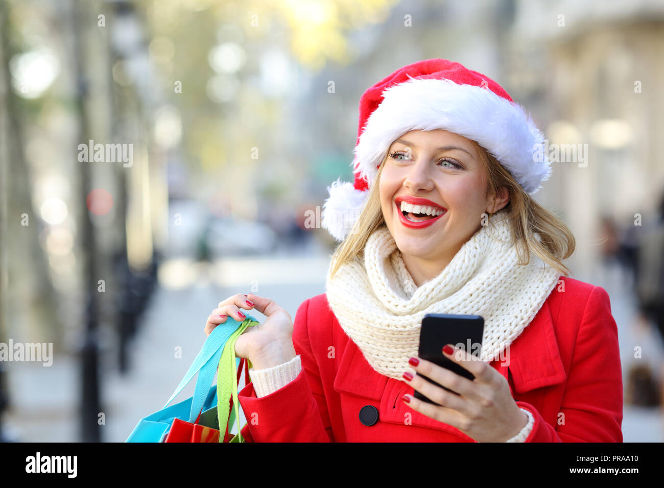 Happy shopper in possesso di un telefono cellulare e shopping bags pensare guardando il lato sul Natale in strada Foto Stock