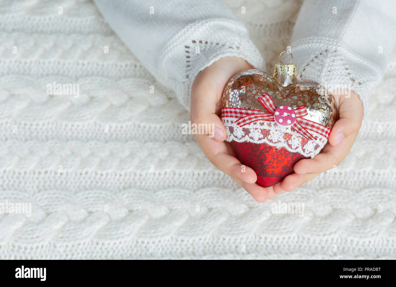 Bambino tiene un cuore di vetro, un giocattolo di Natale nelle mani sullo sfondo di un maglione caldo Foto Stock