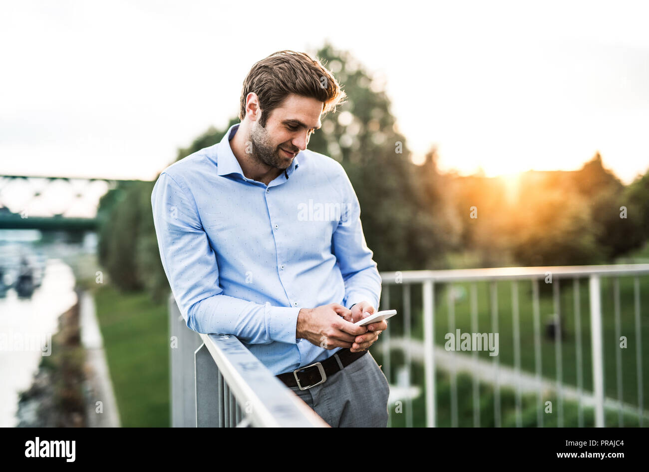 Un giovane imprenditore con lo smartphone in piedi su un ponte al tramonto, poggiando su una ringhiera. Foto Stock