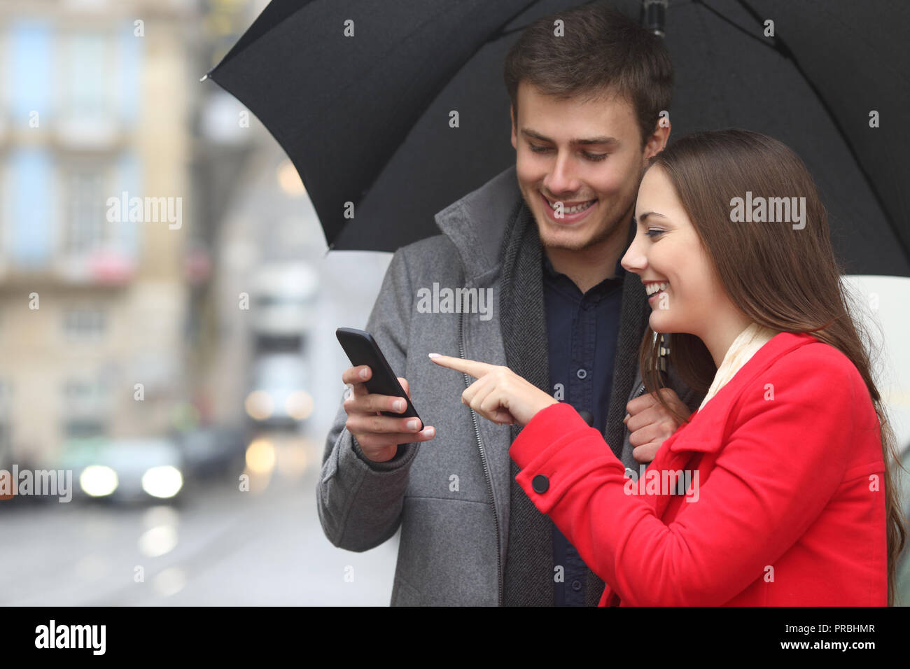 Felice coppia controllo smart phone contenuto con ombrello in inverno sotto la pioggia Foto Stock