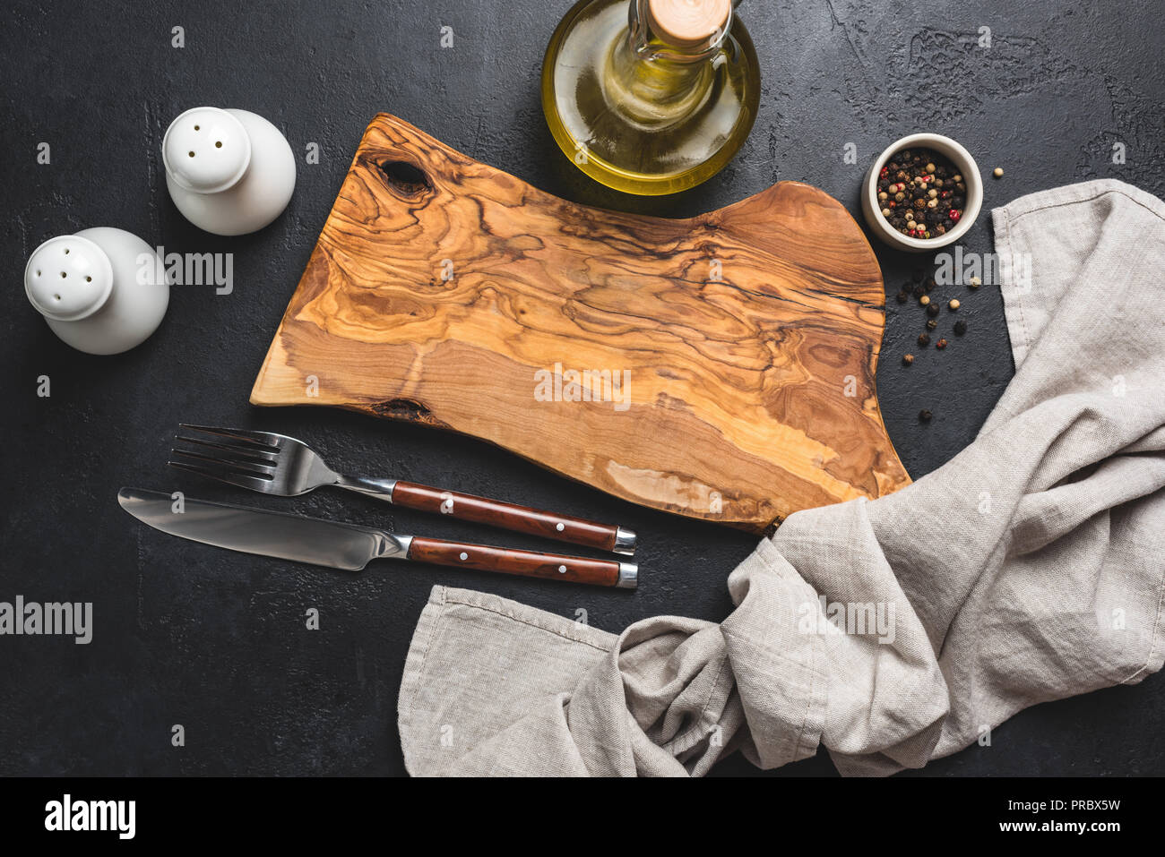 Impostazione tabella nero su sfondo di calcestruzzo. In legno di olivo tagliere, posate, sale e pepe e olio di oliva e biancheria da cucina prodotti tessili Foto Stock