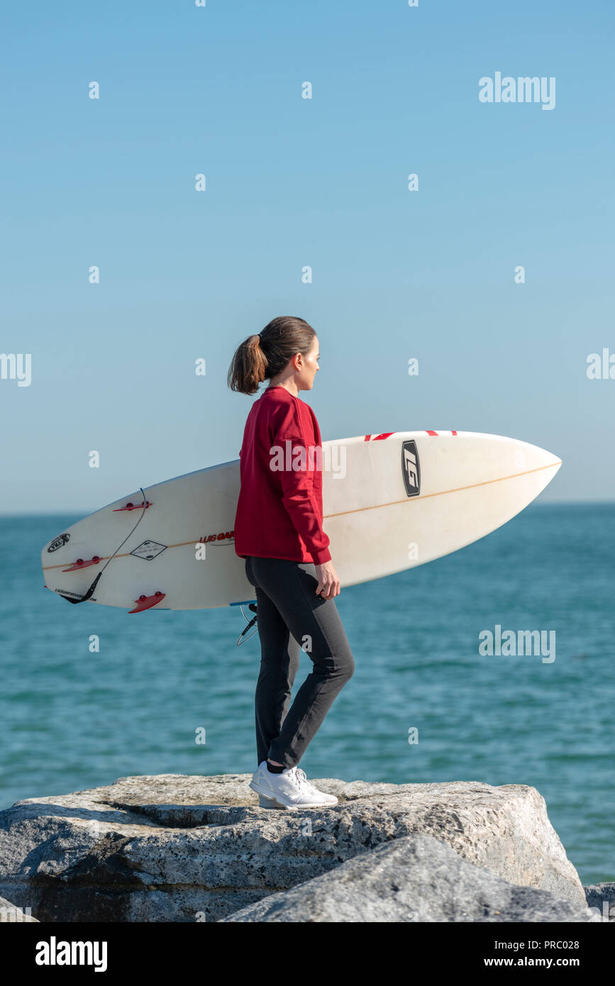 Donna in piedi sulle rocce tenere una tavola da surf controllo del mare prima di surf. Foto Stock