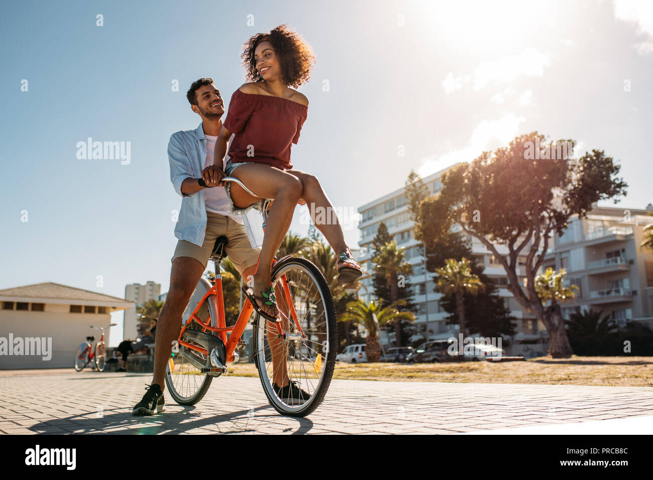 Felice l'uomo e la donna per divertirsi in sella ad una bicicletta su una giornata di sole. Uomo in sella ad una bicicletta nella strada con il suo amico femmina seduti sulla maniglia. Foto Stock
