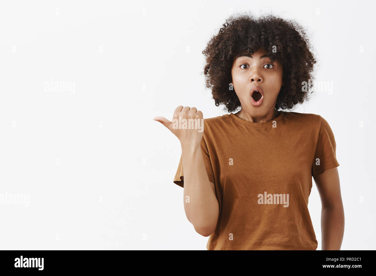 Innalzate eccitato carino di carnagione scura la ragazza con i capelli ricci in marrone alla moda di t-shirt rivolta verso sinistra con il pollice senza fiato e dire wow con piegato aperto bocca essendo sotto impressione domande Foto Stock