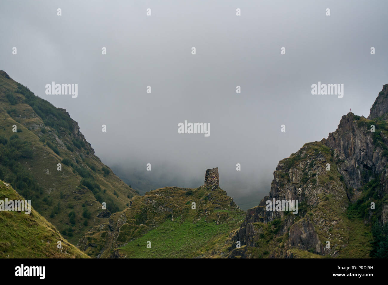 Una solitaria torre in pietra shelter in rovina sorge sulla sommità di una collina rocciosa circondata da grandi montagne rocciose e basso nuvoloso nebbia. Foto Stock