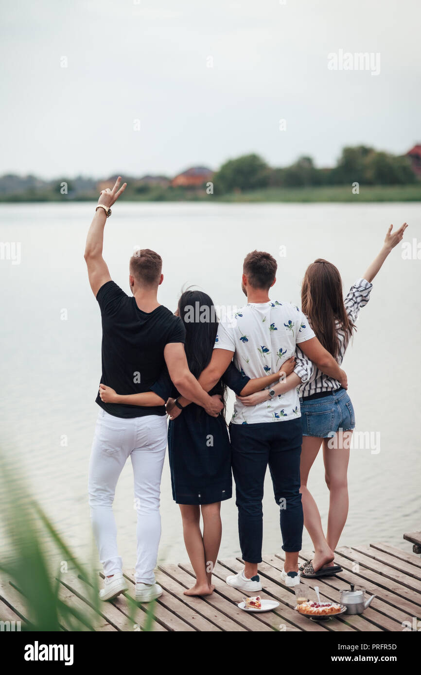 Compagnia di giovani in piedi, guardando il lago, vista posteriore. Amici godere di riposo nella natura, sul molo di legno in fiume Foto Stock