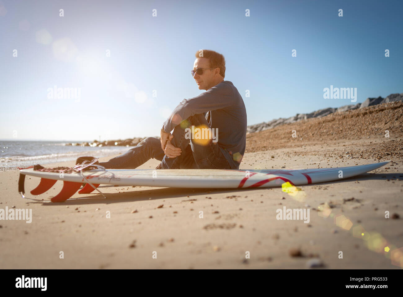 Maschio maturo surfer seduto sulla spiaggia con una tavola da surf a guardare le onde, lens flare Foto Stock