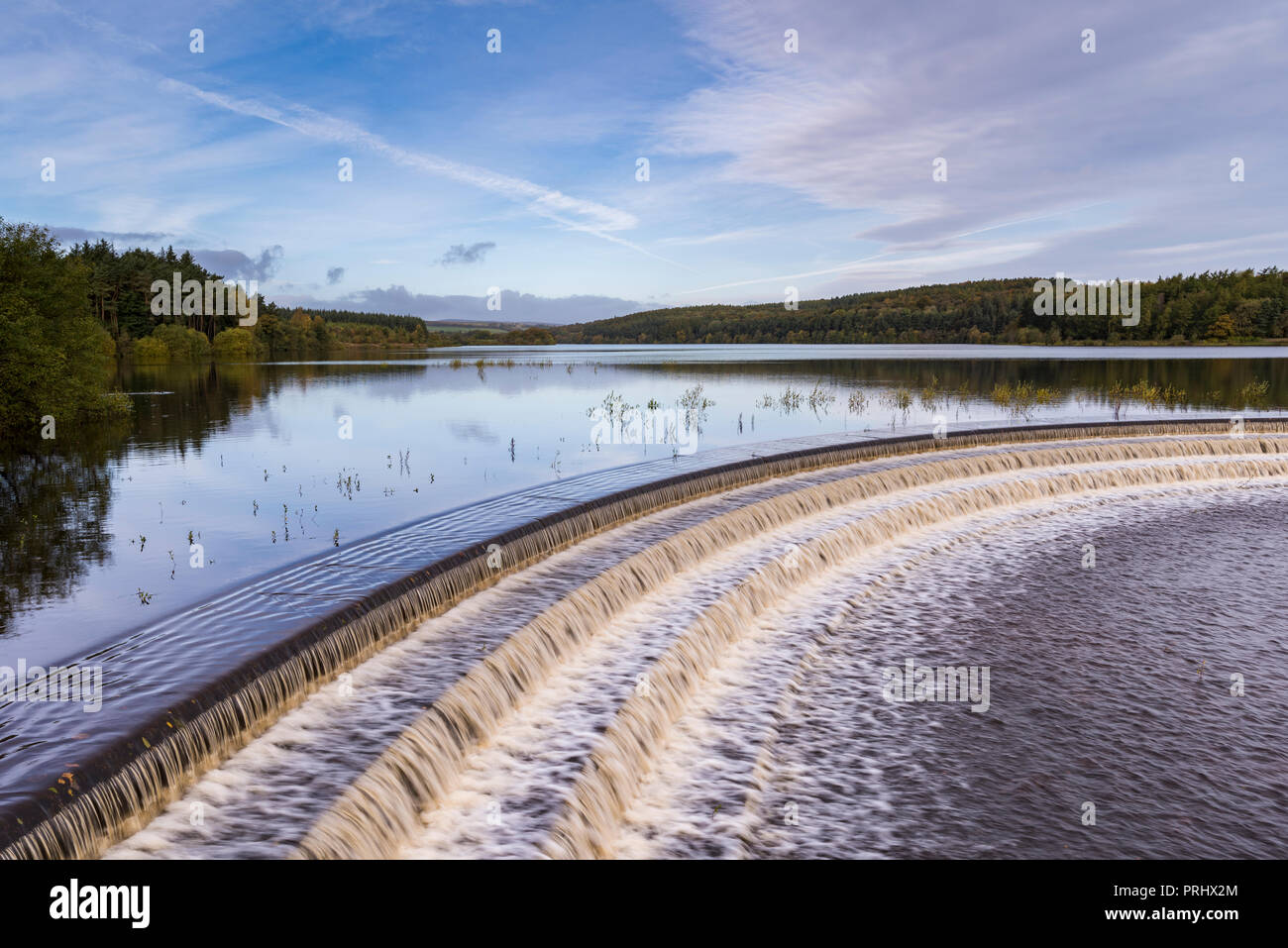 L'acqua che scorre su stramazzo da calme scenic viale alberato di lago, sotto il profondo blu del cielo - Serbatoio Fewston, Washburn Valley, North Yorkshire, Inghilterra, Regno Unito. Foto Stock