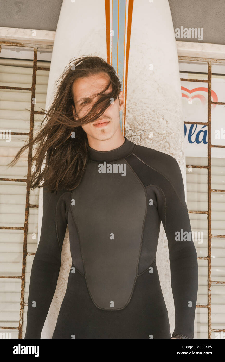 Ritratto di uomo con capelli lunghi in neoprene con il surf board guardando la fotocamera Foto Stock