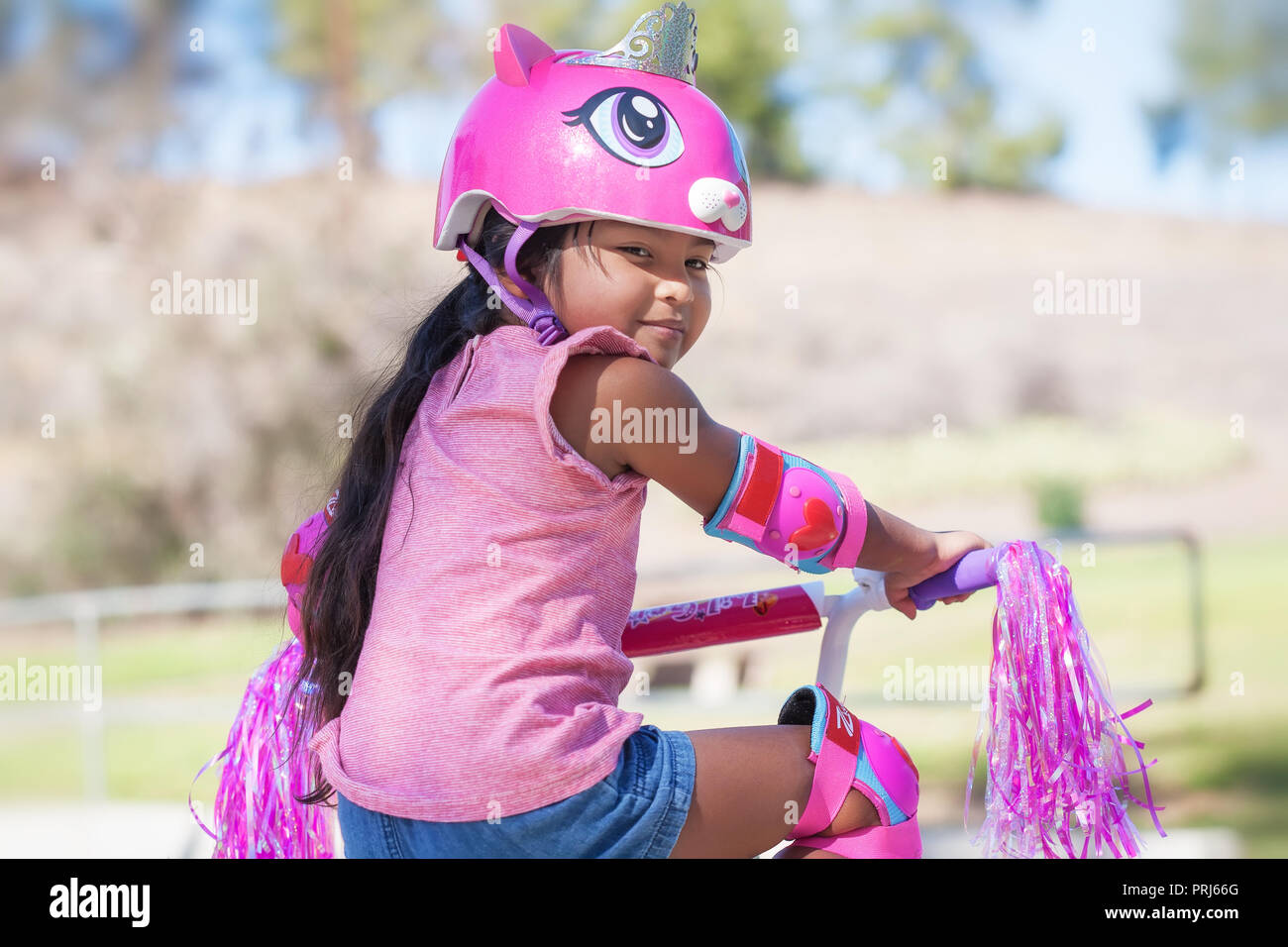5 anno vecchia ragazza ottenere pronto per cavalcare lontano nel suo moto rosa, indossando il casco protettivo e ginocchiere in un parco con le colline Foto Stock