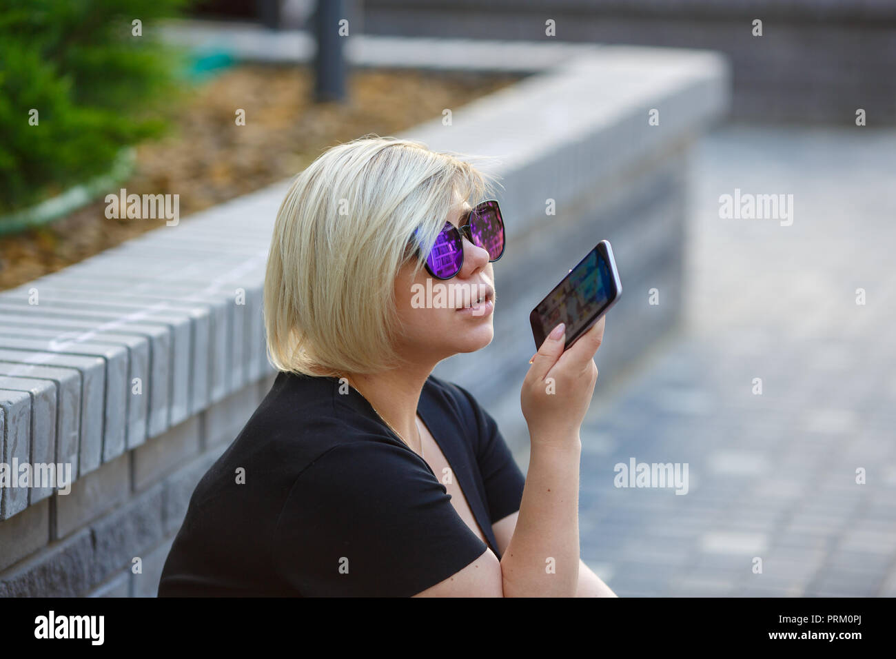 Ritratto ragazza giovane studente in occhiali da sole dimensioni plus si trova nel cortile del campus, parlando al telefono con gli amici, all'aperto, a sinistra di un messaggio audio, vo Foto Stock