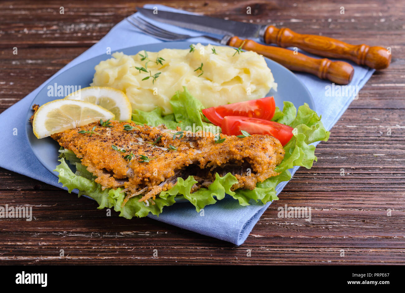 Frittura di pesce piatto su insalata verde con purè di patate, pomodori e le fette di limone in blu sulla piastra scuri in legno rustico sfondo. Deliziosa cena. Foto Stock