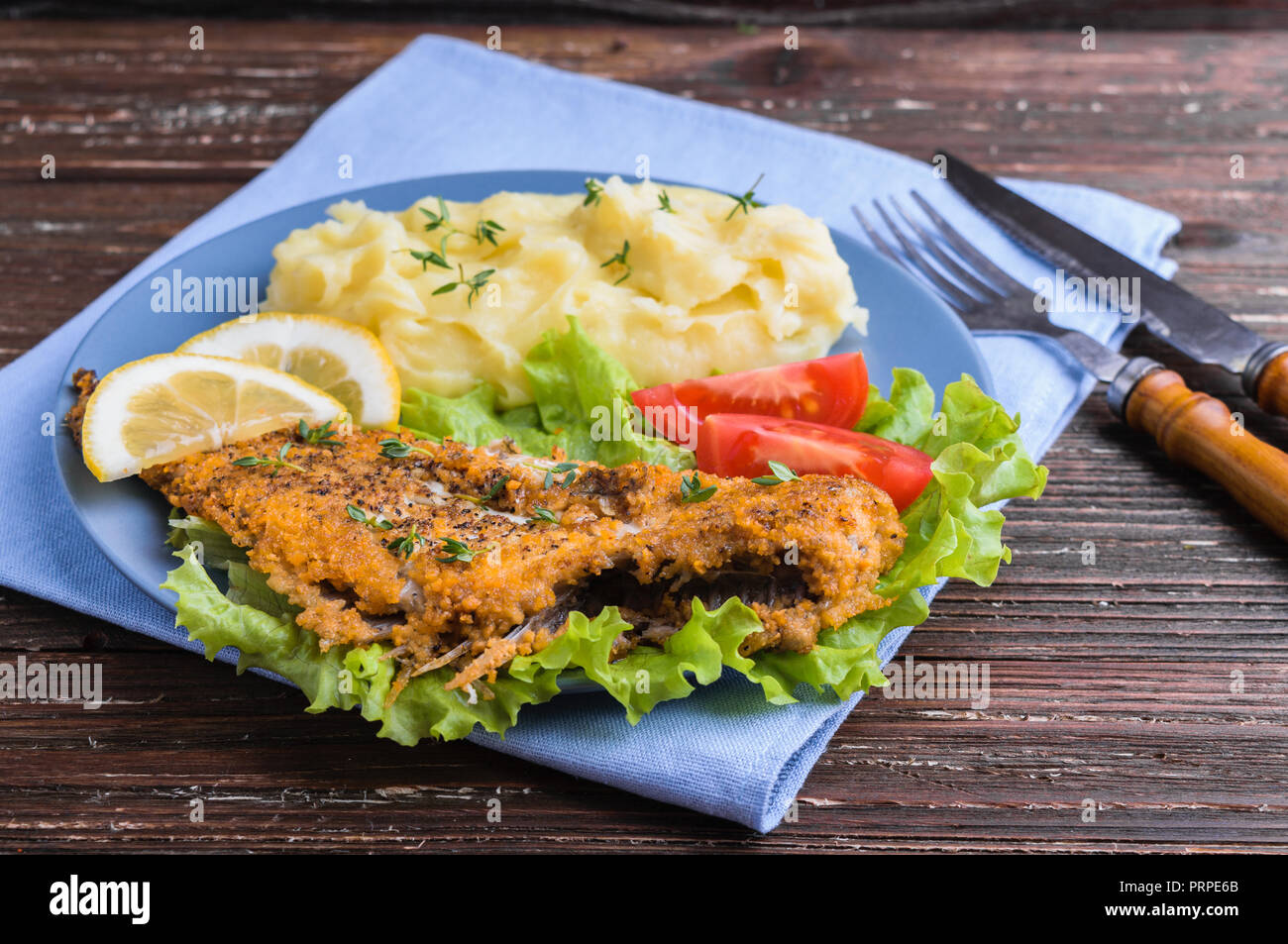 Frittura di pesce piatto su insalata verde con purè di patate, pomodori e le fette di limone in blu sulla piastra scuri in legno rustico sfondo. Deliziosa cena. Foto Stock