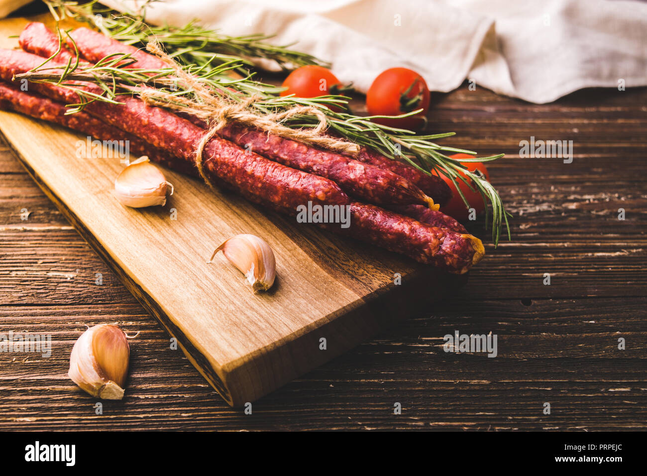 Kabanosy - affumicato polacca tradizionale salsiccia di maiale sul tagliere di legno. Con erbe di rosmarino, spicchi di aglio e i pomodori. Su tavola in legno rustico Foto Stock