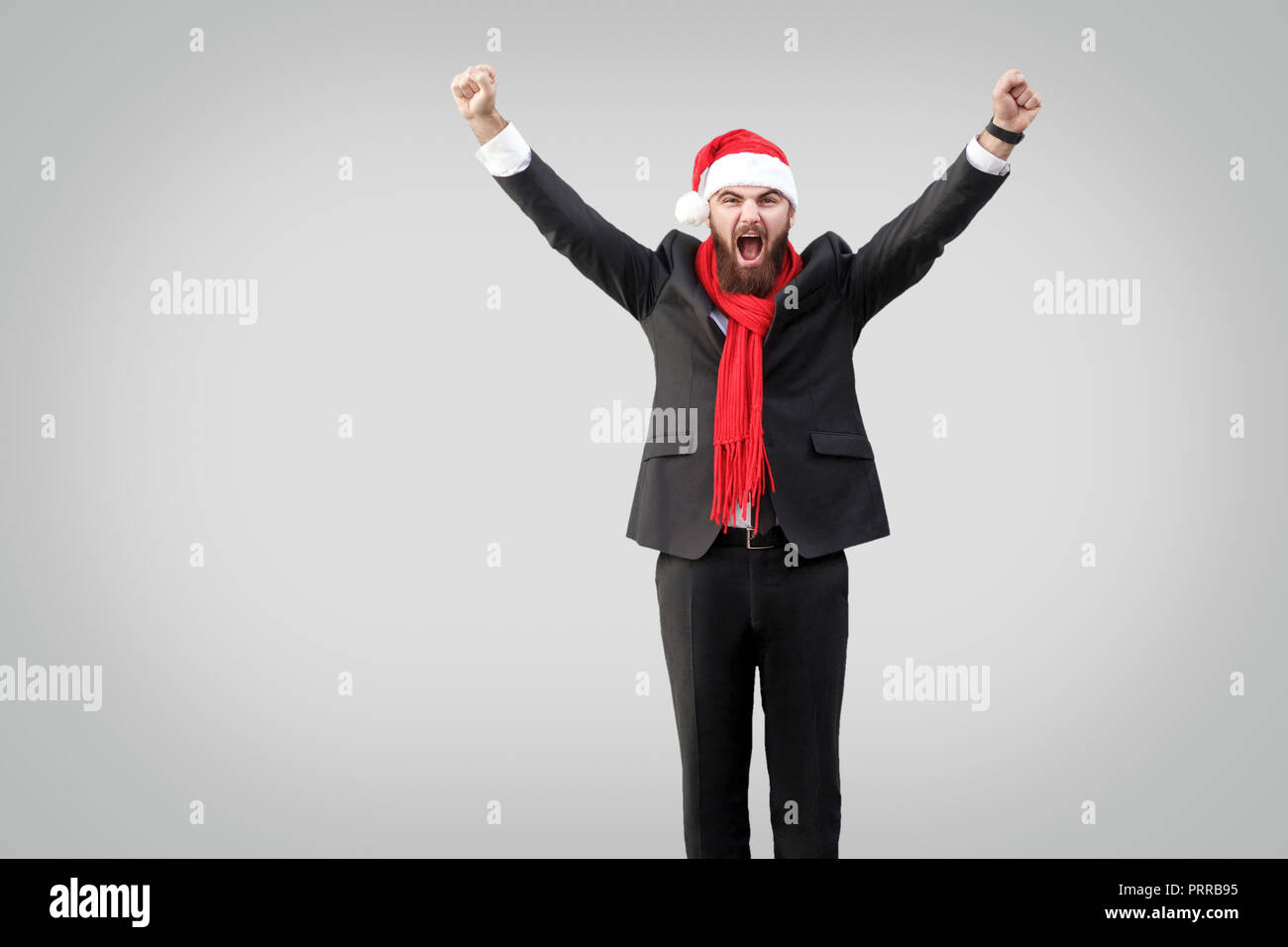 Imprenditore barbuto in classico abito nero, sciarpa rossa e nuovo anno hat, guardando la telecamera con i bracci sollevati e urlando. Wining e concetto di successo. Ho Foto Stock