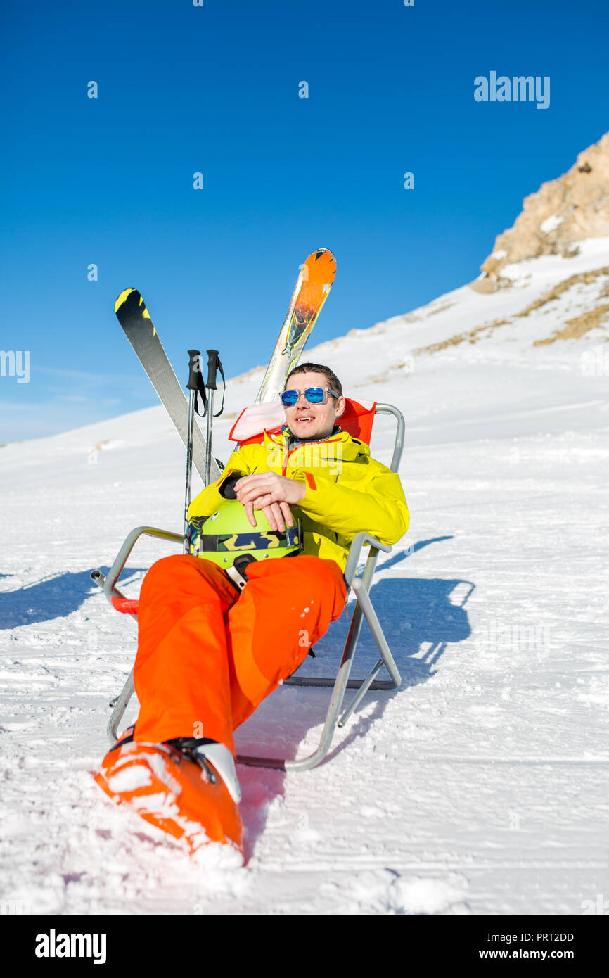 Immagine di sport uomo seduto sulla sedia accanto a sci e bastoni sullo sfondo di montagne innevate Foto Stock