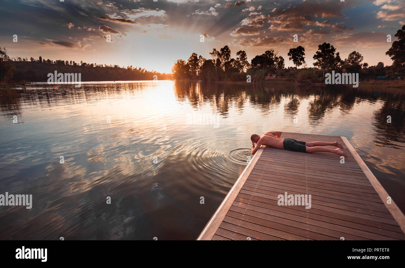 Giovane uomo senza maglietta sdraiato a faccia in giù sul pontile in legno bagnando la sua testa in acqua di lago Foto Stock