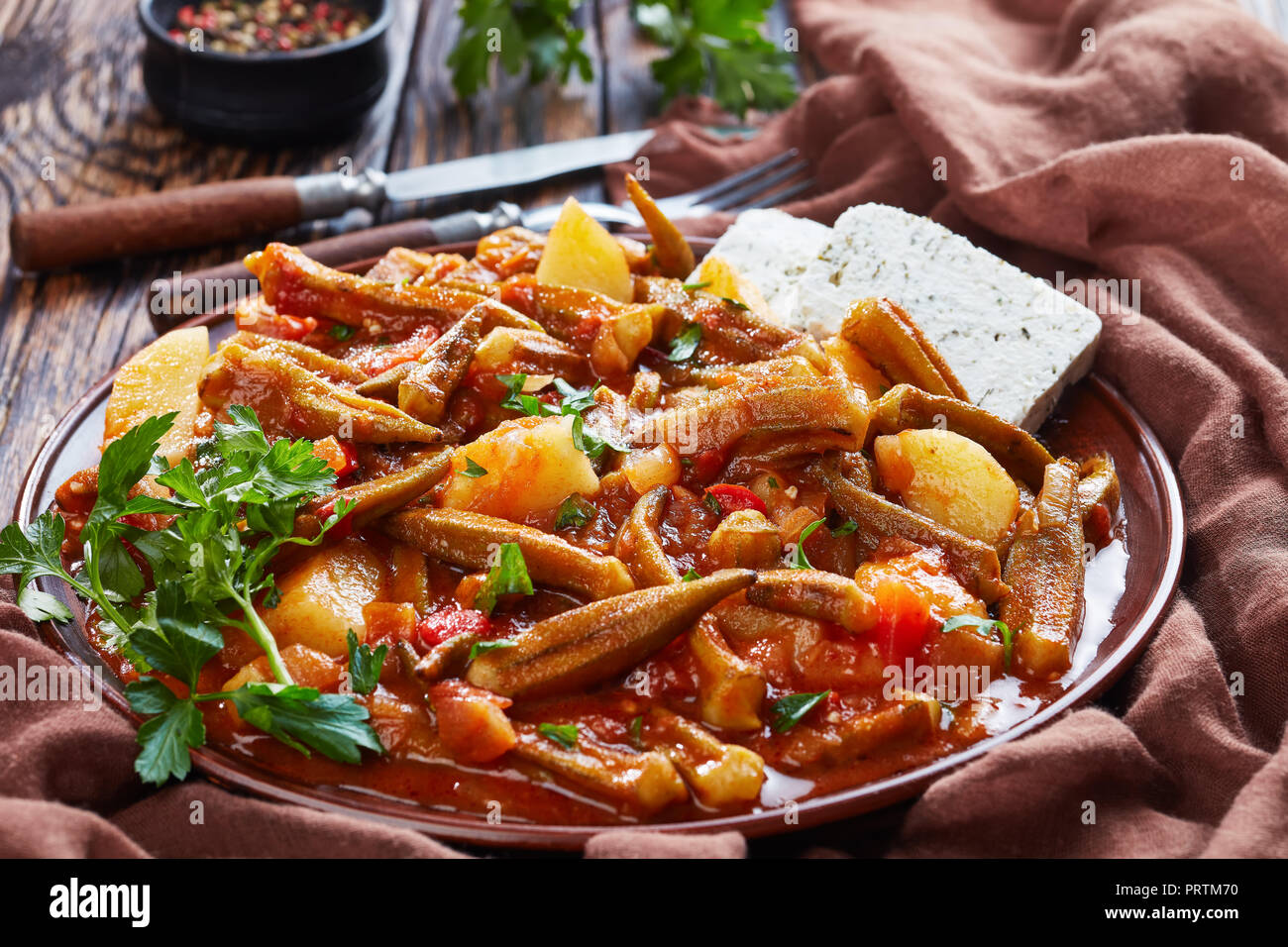 Stufati di okra con pomodoro e patate servita con il formaggio feta le fette su un piatto di terracotta su un tavolo rustico, ricetta greca. forcella, coltello, prezzemolo al Foto Stock