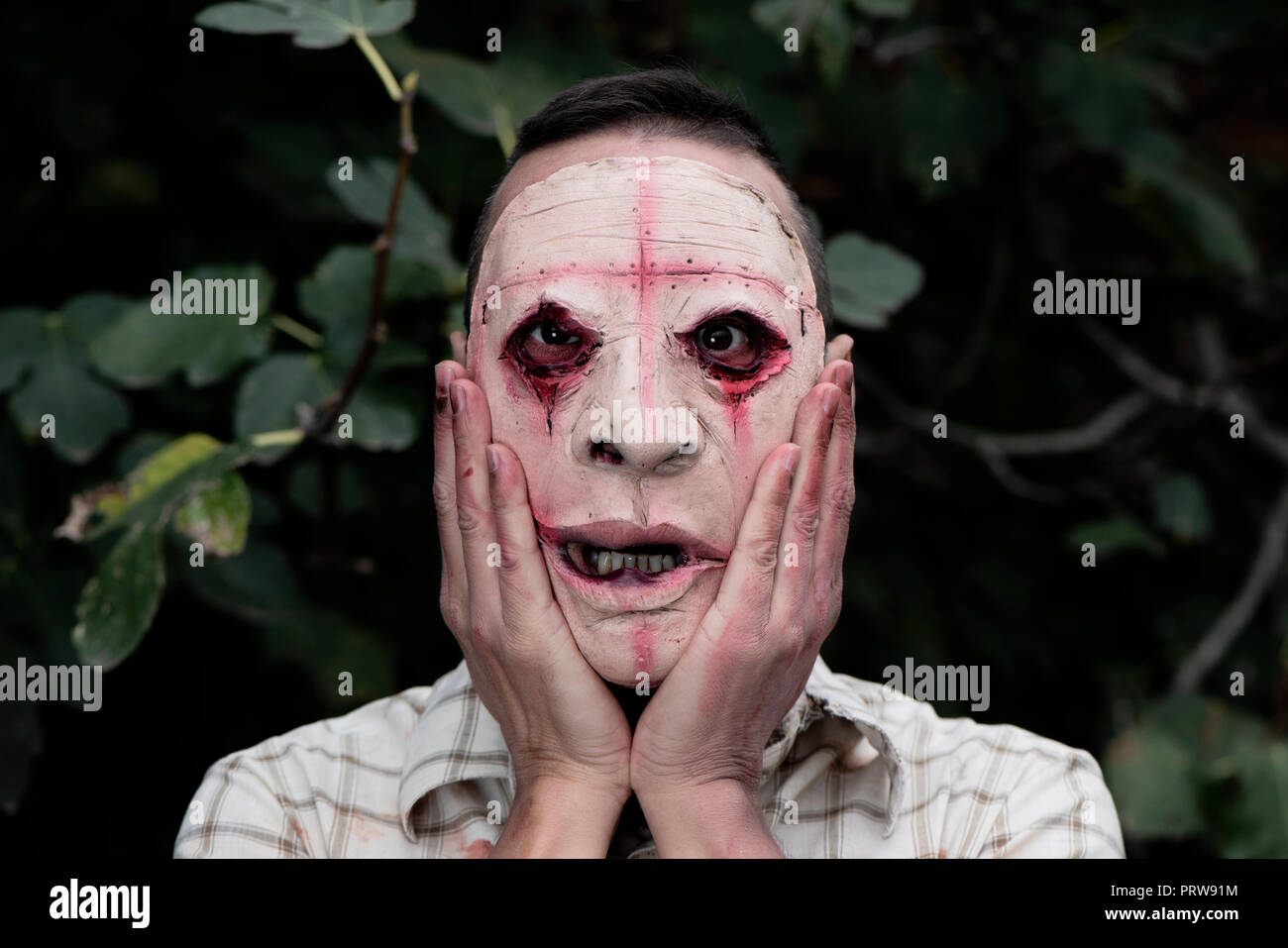 Primo piano di un pauroso sfigurato uomo, indossando sporchi e logori vestiti con le sue mani nel suo volto, nei boschi Foto Stock