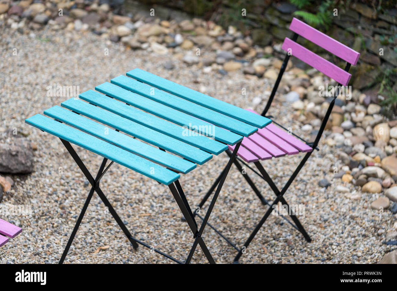 Una rosa di legno sedia pieghevole e un quadrato tavolo blu sono in piedi sulla ghiaia nel cortile di una caffetteria nella luce naturale di sera Foto Stock