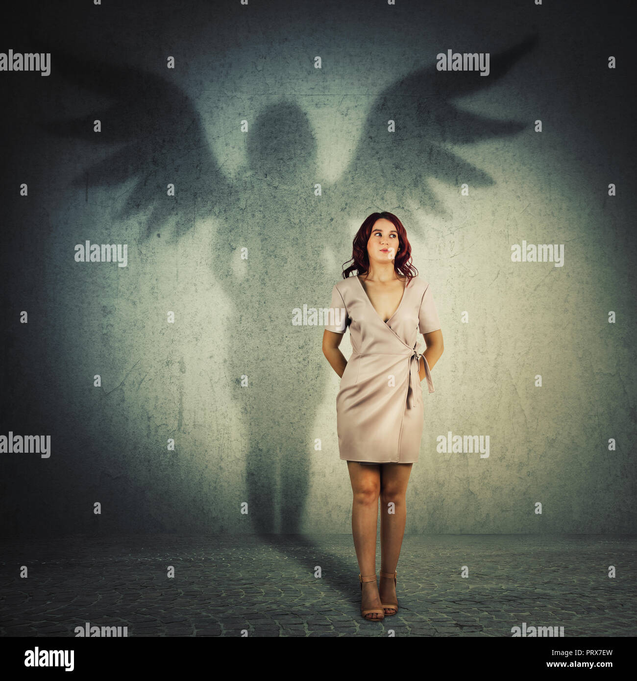 Funny giovane donna nascondendo le mani dietro la schiena che guarda lontano. A piena lunghezza ritratto femminile colata di un supereroe ombra con angelo ali in una stanza buia wa Foto Stock