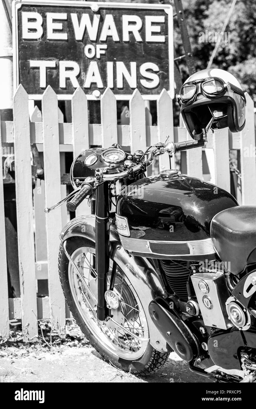 Vista laterale in bianco e nero ravvicinata, Vellocette vintage moto, casco, googles, parcheggiato da picchetto recinto presso la stazione ferroviaria Heritage, evento anni '40. Foto Stock