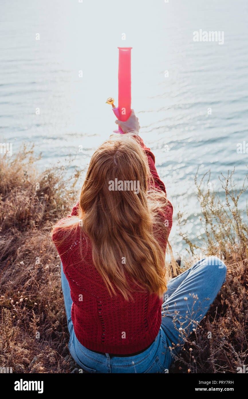 Una giovane ragazza ribelle in un maglione rosso di fumare su una costa rocciosa vicino al mare nella golden sunrise. Foto Stock