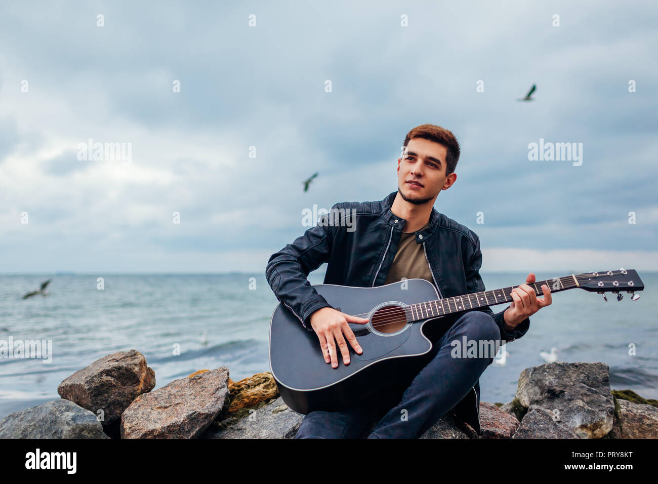 Giovane uomo con la chitarra acustica a suonare e cantare da solo sulla  spiaggia circondata da rocce sul giorno di pioggia Foto stock - Alamy