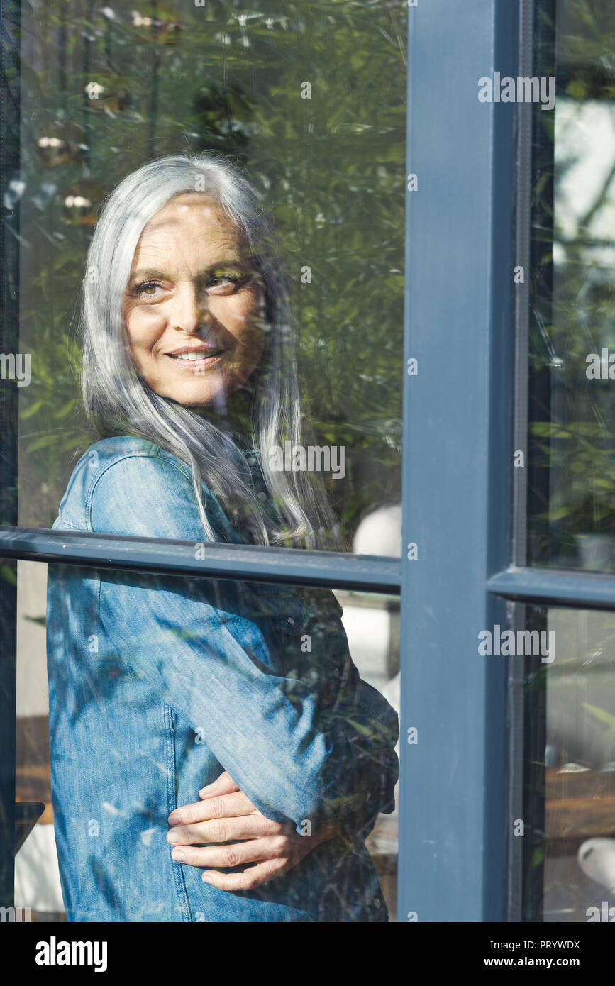 Senior donna guardando fuori della finestra, sorridente Foto Stock