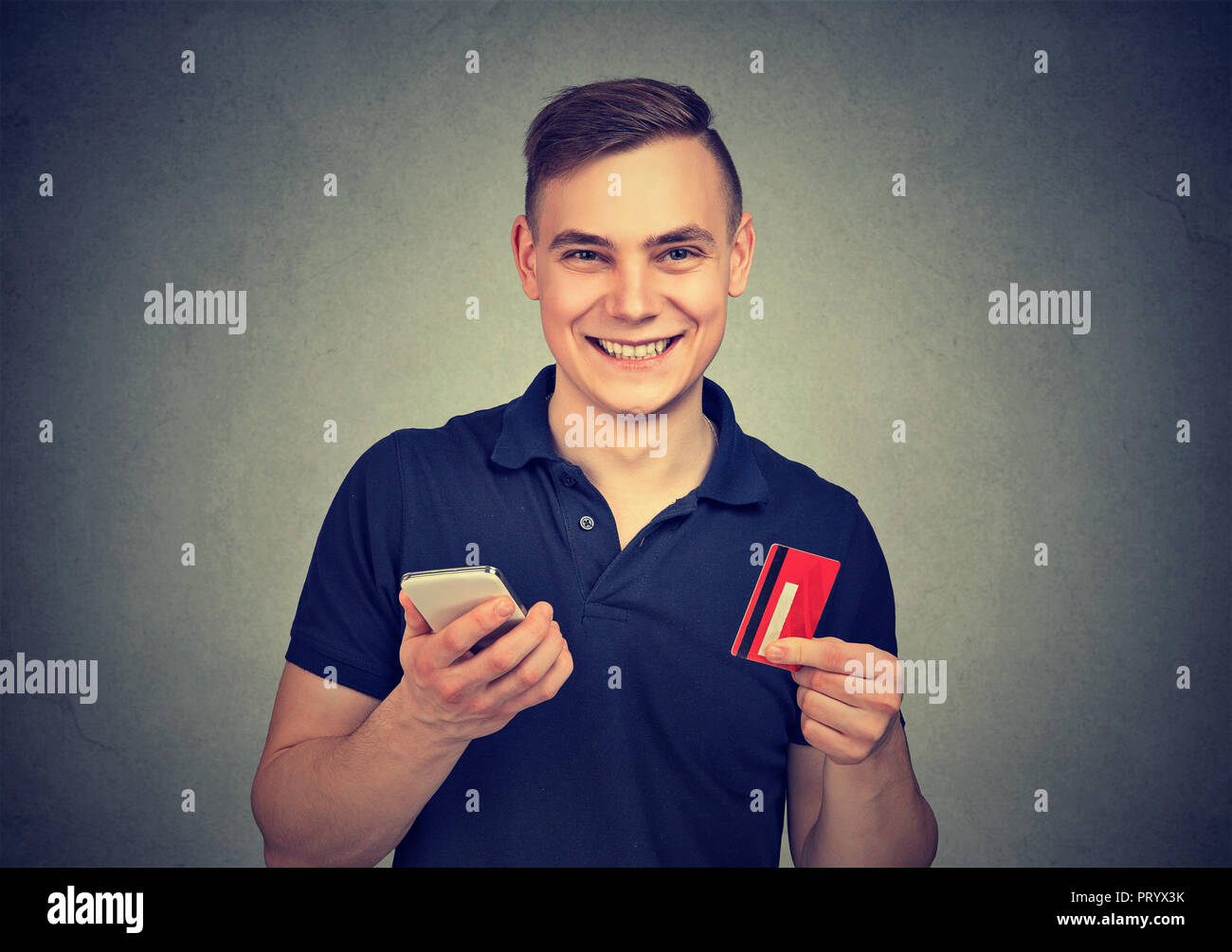 Bel giovane uomo con carta di credito e il telefono cellulare è eccitato con servizio comodo e sorridente in telecamera Foto Stock