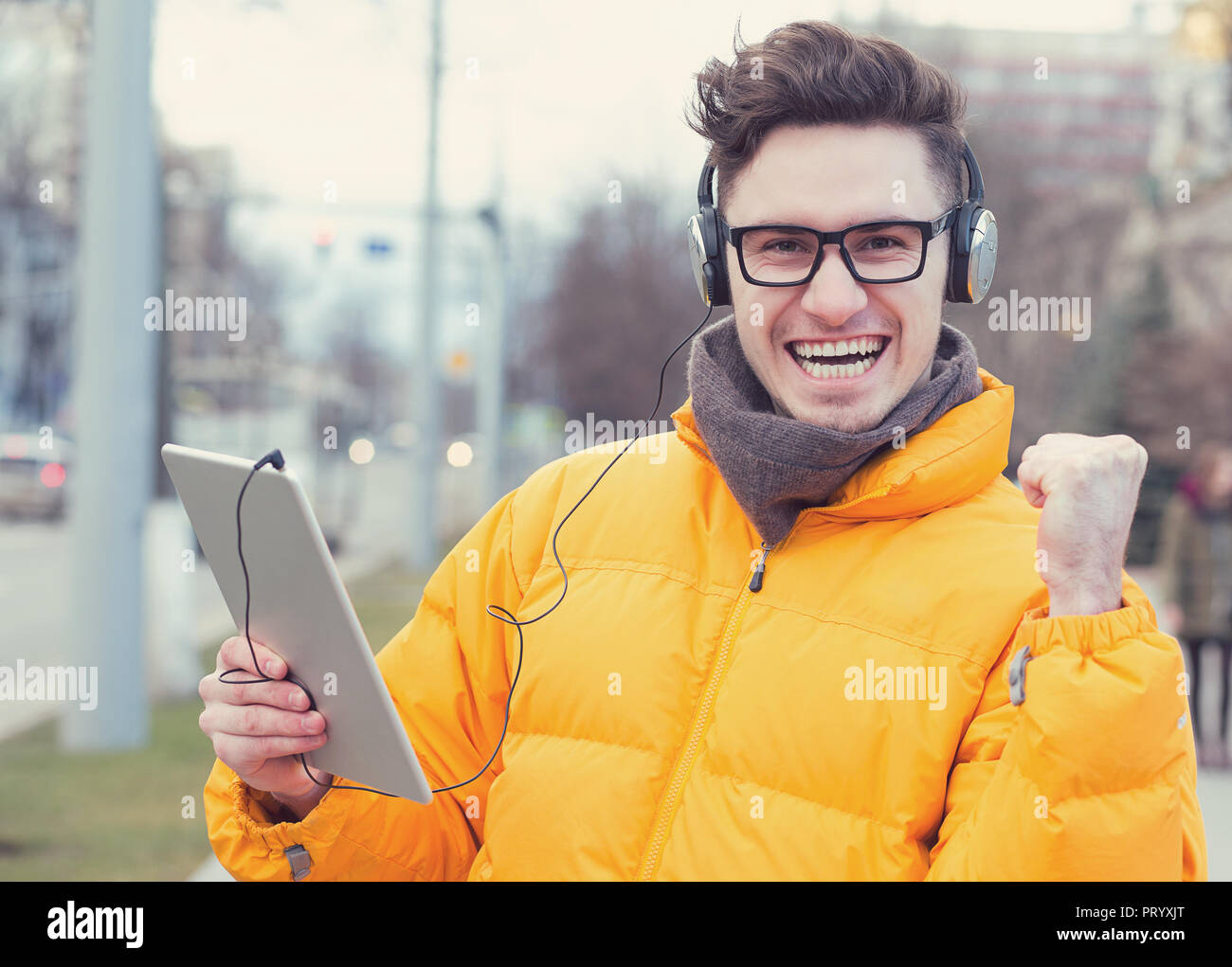Felice l'uomo di successo nel mantello e cuffie holding tablet e pugno fino sulla strada eccitato con win Foto Stock