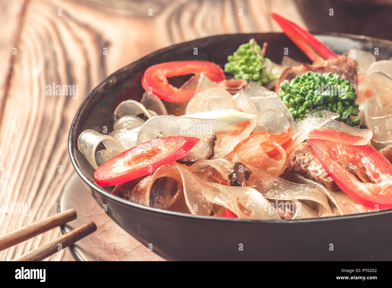 Hot asian piatto piccante tagliato spesso tagliatelle di vetro Mung Bean fungosa in agrodolce carne in salsa di peperoni verdure broccoli porro in ciotola bacchette sul tavolo da cucina di salsa di soia. Foto Stock