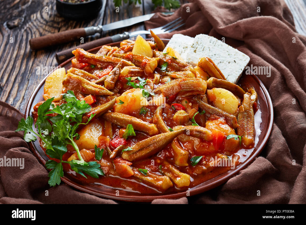 Stufati di okra con pomodoro e patate servita con il formaggio feta le fette su un piatto di terracotta su un tavolo rustico, ricetta greca. forcella, coltello, prezzemolo al Foto Stock