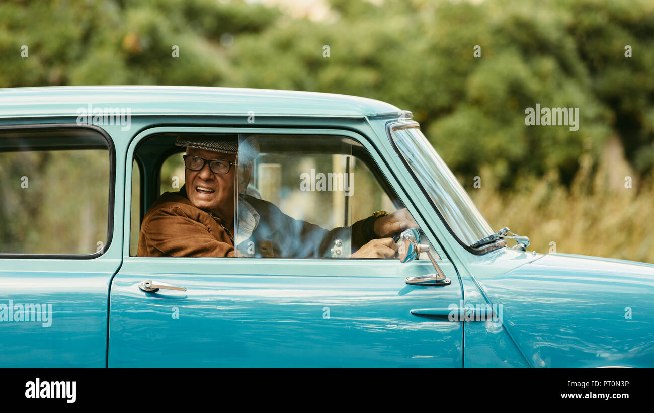 Senior uomo alla guida di un auto classica. Il vecchio uomo guarda fuori dalla finestra della sua vettura prima di fare un giro su strada. Foto Stock