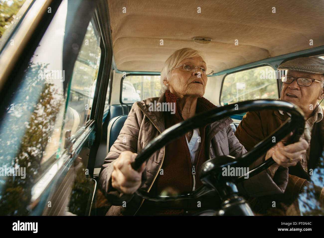Il vecchio donna alla guida di una vettura e parlare con suo marito in seduta sedile passeggero. Coppia di anziani la guida in auto d'epoca. Foto Stock