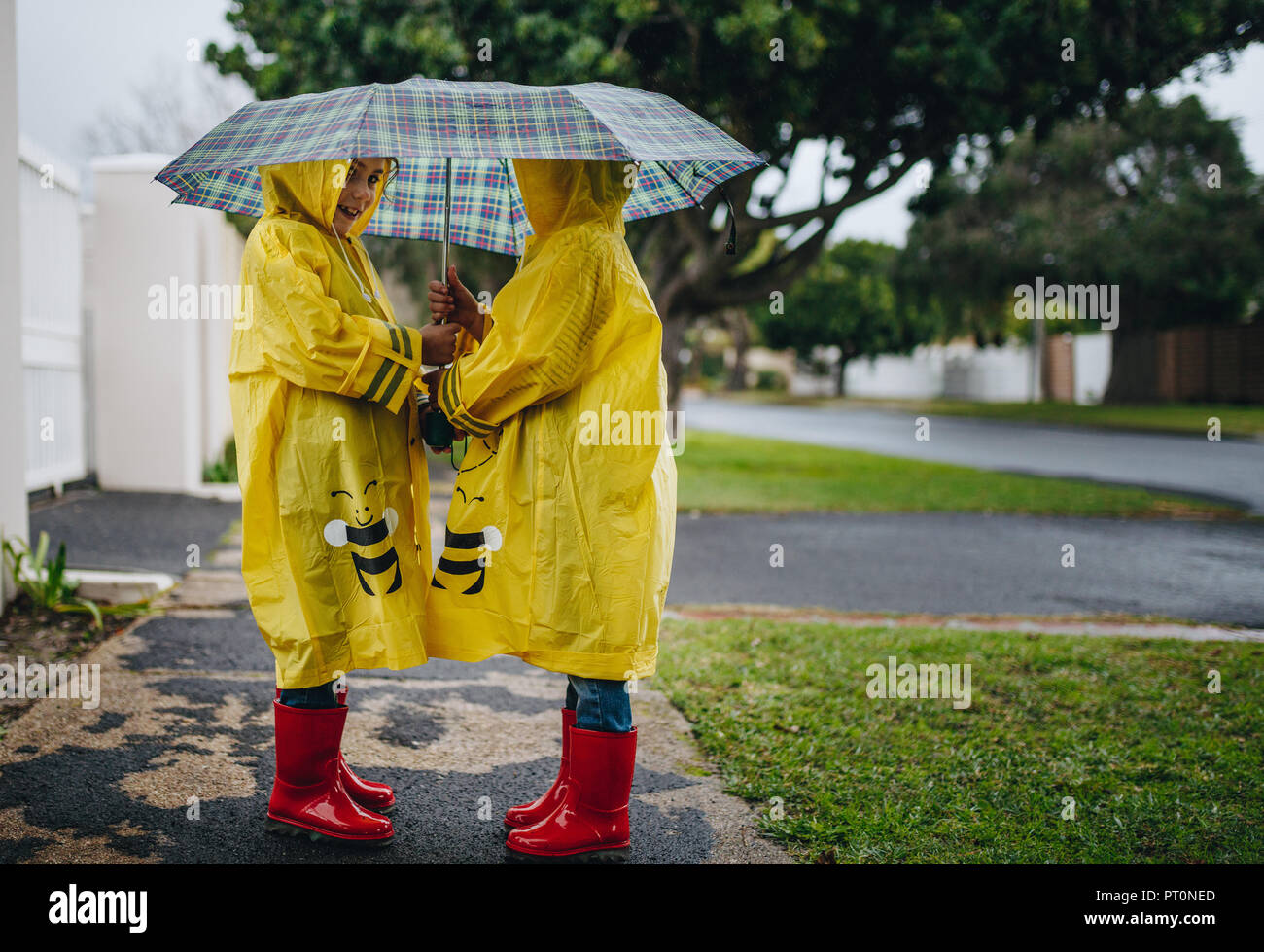 I due ragazzi in IMPERMEABILI CAPPOTTI rosso e stivali di gomma in piedi  sotto l'ombrello. Bambine all'aperto in un giorno di pioggia Foto stock -  Alamy