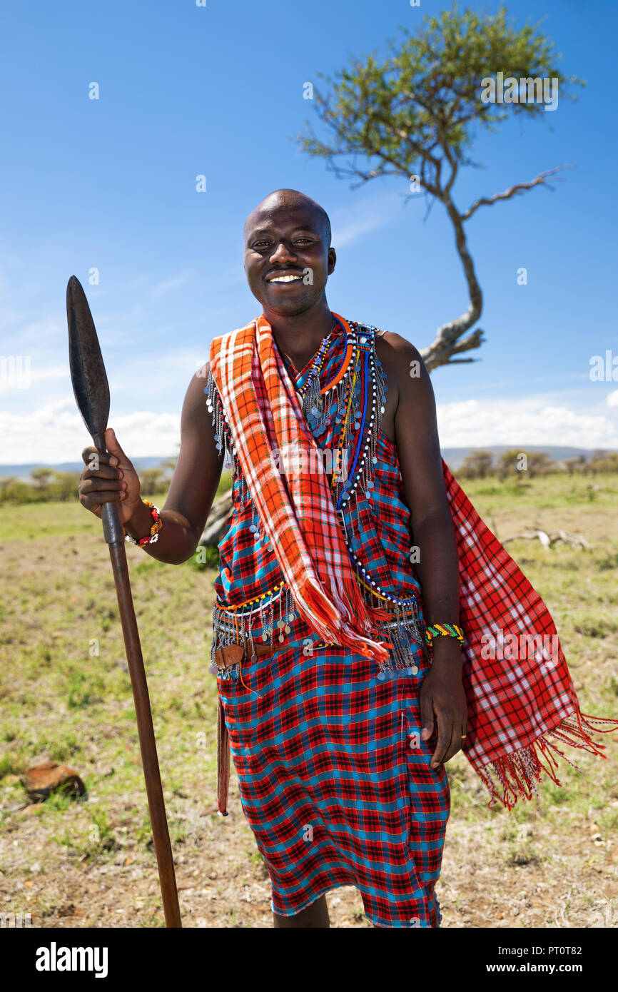 Il Masai Mara riserva nazionale, Kenya, Africa: sorridente giovane guida Maasai con tradizionale in rosso-verifica di vestire e lancia con sfocato Foto Stock