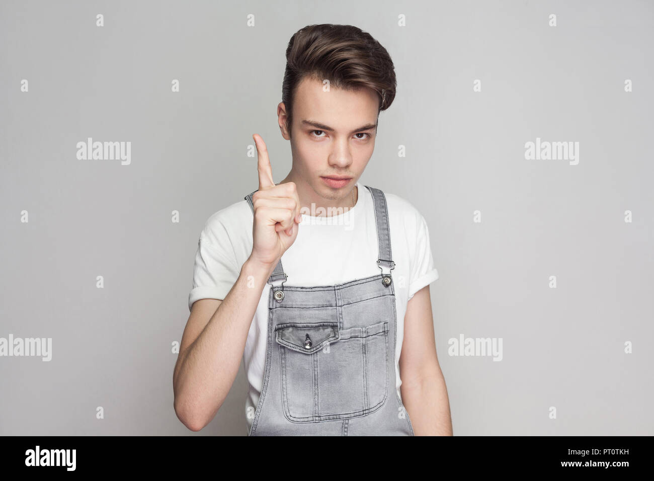 Ritratto di giovane serio brunette uomo in stile casual con t-shirt bianco jeans e tute da lavoro in piedi e guardando la telecamera con il segnale di avvertimento. interna Foto Stock