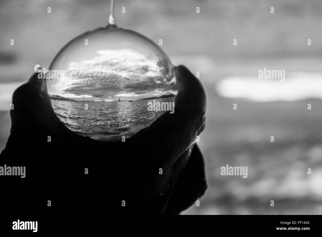 Argento e grigio sole e nuvole seascape con piccola barca sull orizzonte catturato in astratto Palla di vetro tenuto in mano con gocce d'acqua Foto Stock