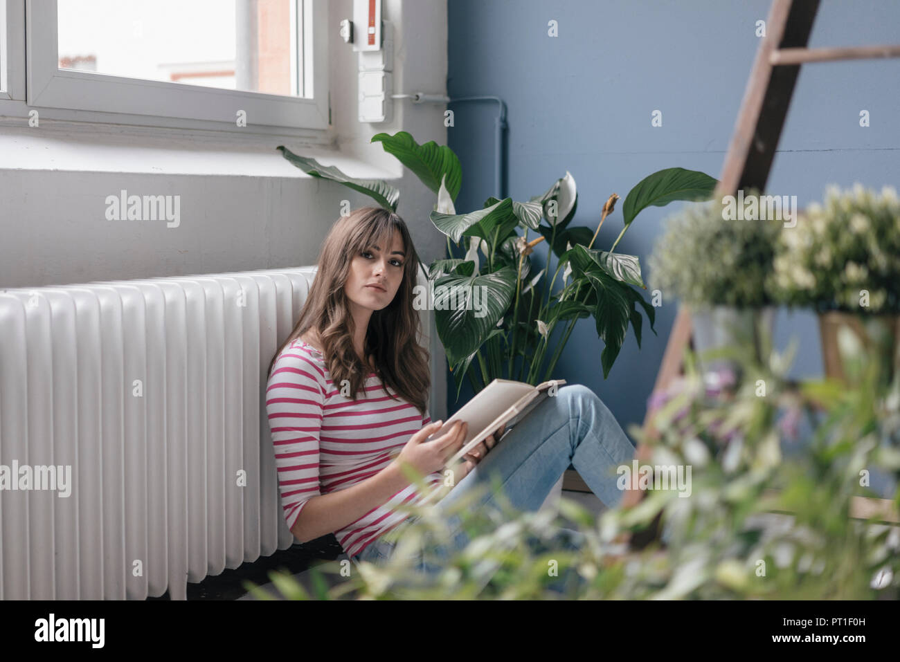 Donna seduta su un terreno nella sua nuova casa, la lettura di un libro, circondato da piante Foto Stock