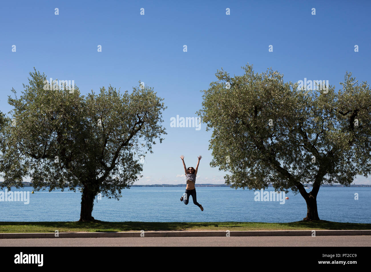 L'Italia, il Lago di Garda, felice giovane donna di saltare in aria tra due alberi Foto Stock